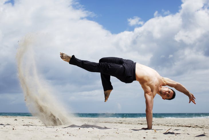 Capoeira instructor posing in a move while kicking up sand on a beach