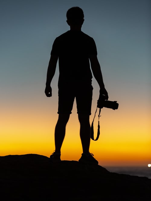 Silhouette of a wildlife photographer holding a camera and looking out at the sunset over the ocean